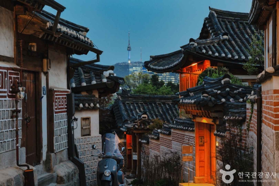 A steep hanok alley with tiled eaves overhead and N Seoul Tower visible between the roofs
