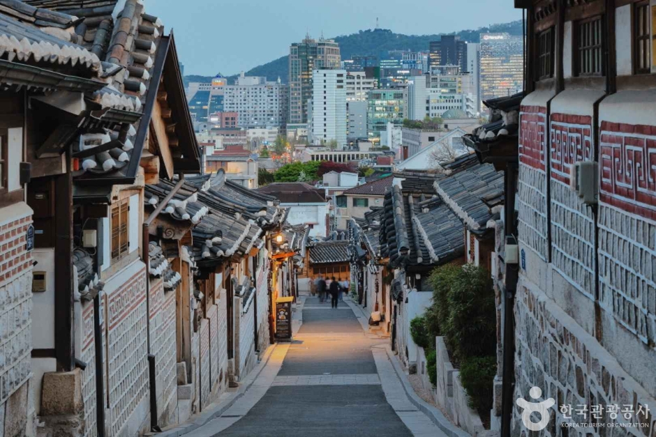 A narrow sloping lane between hanok houses in Bukchon, with modern Seoul towers rising behind the rooftops