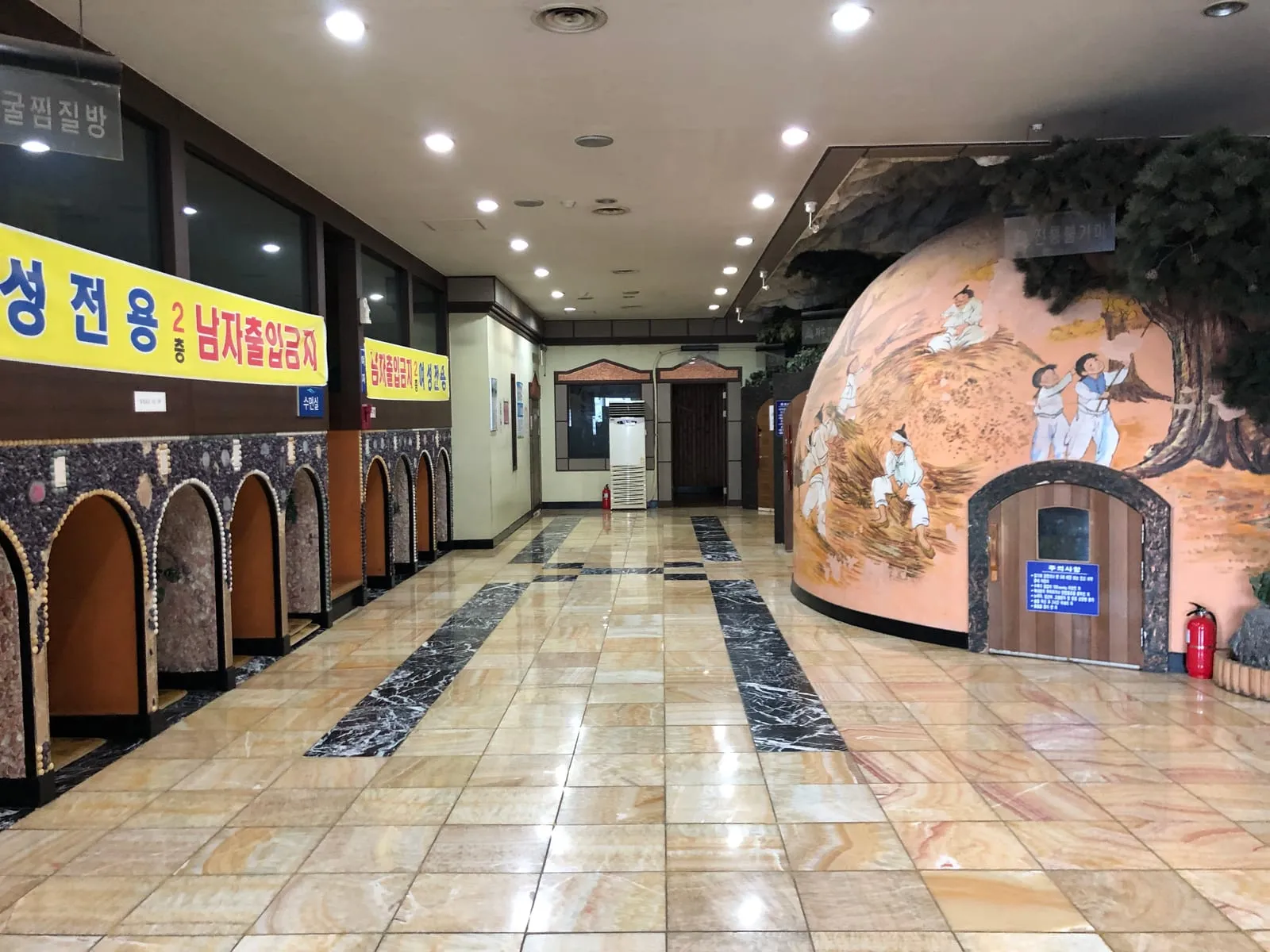 A domed traditional kiln room inside a Korean sauna, painted with a folk mural and entered through a small low wooden door, with a row of arched alcoves along the opposite wall