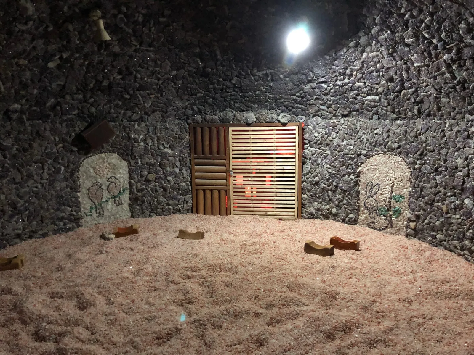 Inside a domed salt room: a floor of coarse pink salt crystals with wooden pillows lying on it, walls of dark crushed stone, and a heater glowing red behind wooden slats
