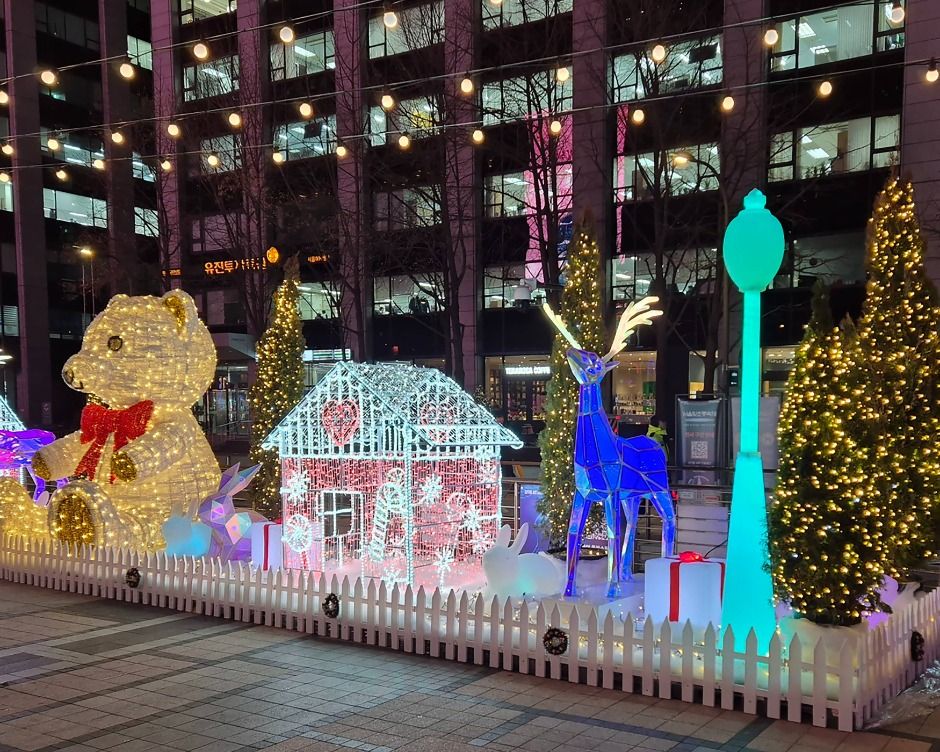 Illuminated animal and house figures behind a low white fence on paving beside a Seoul street at night in winter