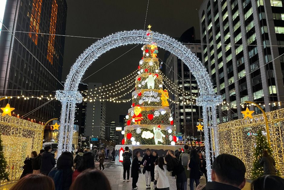 A crowd walking beneath a large illuminated arch toward a lit decorated tower at Cheonggye Plaza in Seoul at night