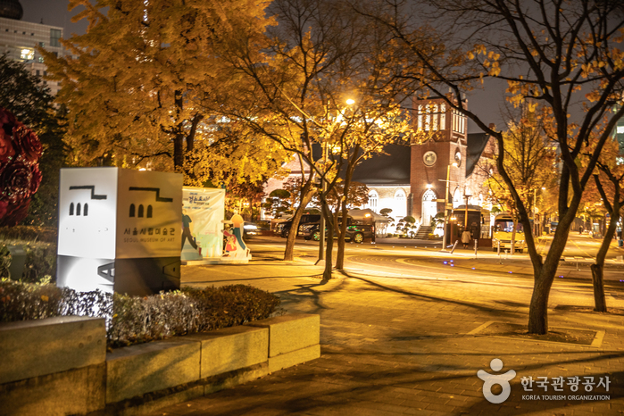 The stone sign of the Seoul Museum of Art at night, with ginkgo trees lit yellow by street lamps and a brick church tower in the distance