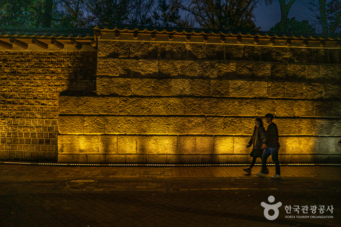 Two people walking past a high wall of large stone blocks at night, lit from below by a row of warm ground lights