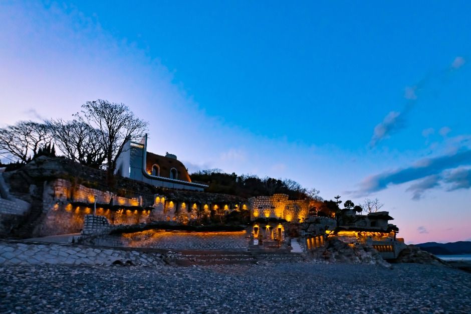 A stone fortress structure on a rocky shoreline lit at dusk, the sea and sky deep blue behind