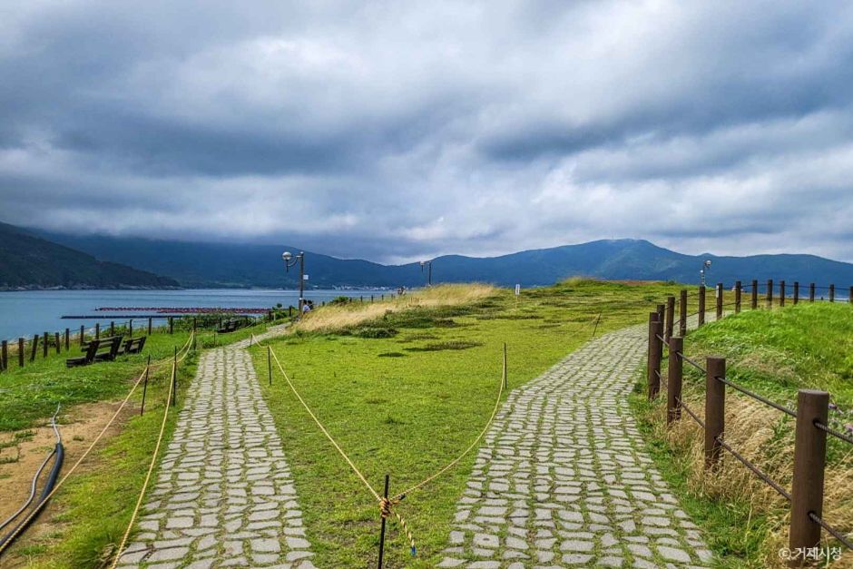 A stone-paved path running along a green headland above a calm blue sea