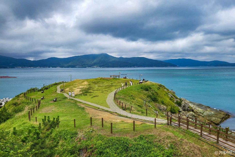 A grassy headland on Geoje island with a footpath curving over it, turquoise sea and low mountains beyond