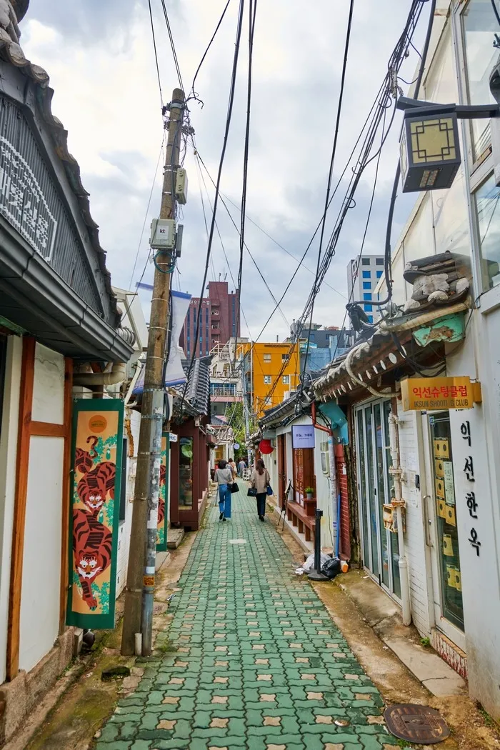 A narrow Ikseondong alley with a vertical sign reading 익선한옥 on a white wall and a yellow sign above it, green tiled path between hanok buildings