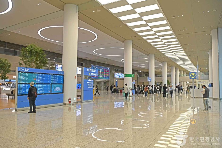 仁川国際空港の到着ロビーを、案内モニターの列の前を通って歩く旅行者たち