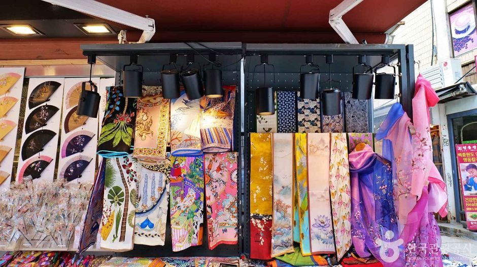 A street stall front in Insadong hung with printed silk scarves, boxed folding fans displayed on the left and hair ornaments in a case below