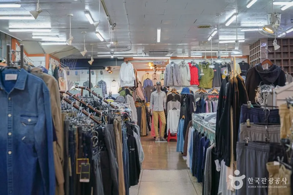 One person walking alone down the aisle of a covered clothing market, racks of shirts and coats on both sides