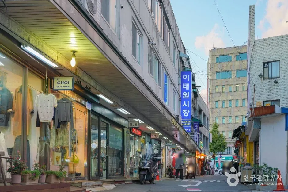夕暮れの梨泰院の路地。片側に明かりのついた店が並び、青い縦看板に「이태원시장 ITAEWON MARKET」とある