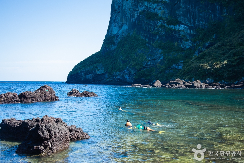 Clear shallow water over dark volcanic reef at Seongsanpo on Jeju, with a steep headland rising across the bay and a few people in the water