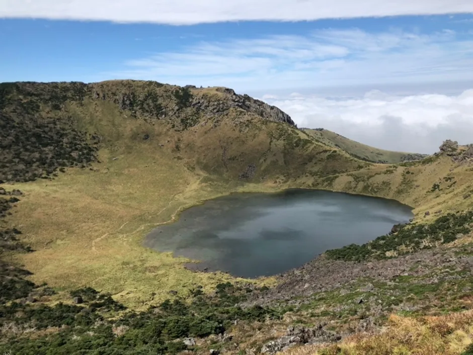 The Baengnokdam crater lake at the summit of Hallasan on Jeju, a shallow pool of water inside a wide green bowl of volcanic rock, cloud lying below the rim
