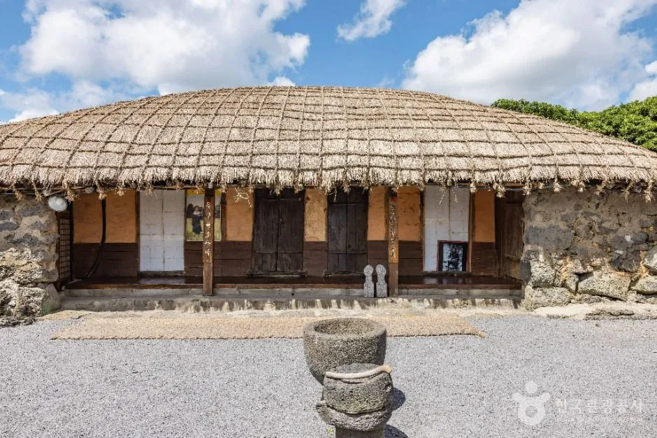 A thatched Jeju house at Seongeup Folk Village, its reed roof bound down under a grid of thick rope, with wooden doors, a low stone base and a stone mortar standing in the swept yard