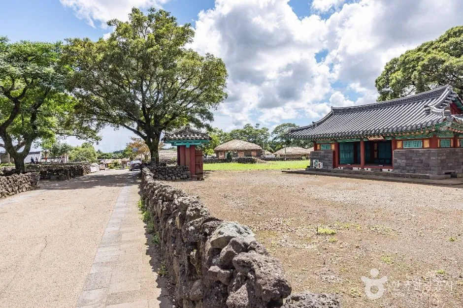 A lane at Seongeup Folk Village on Jeju running between loosely stacked black basalt walls, curving toward a tiled gateway with trees behind
