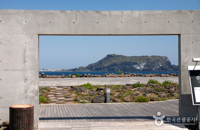 A concrete rectangular opening in a wall, framing Seongsan Ilchulbong across the water, with a wooden deck floor, black volcanic gravel and low planting in front of it