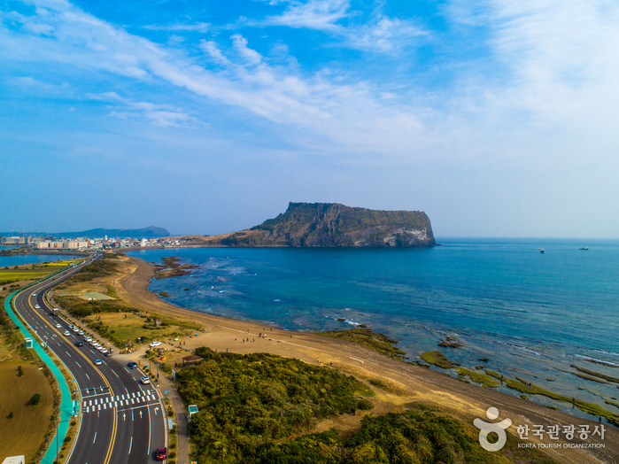 A curving coastal road with a painted cycle lane running above a pale sand beach, with the flat-topped Seongsan Ilchulbong tuff cone rising across the water in the middle distance under a blue sky