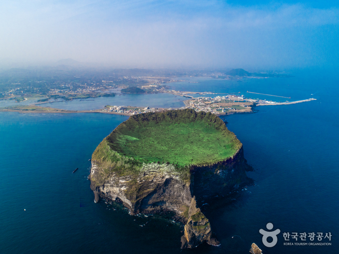 Aerial view of Seongsan Ilchulbong, a cliff-walled tuff cone with a green grass bowl in its crater, joined to the island of Jeju by a narrow sand isthmus with a village and harbour behind it