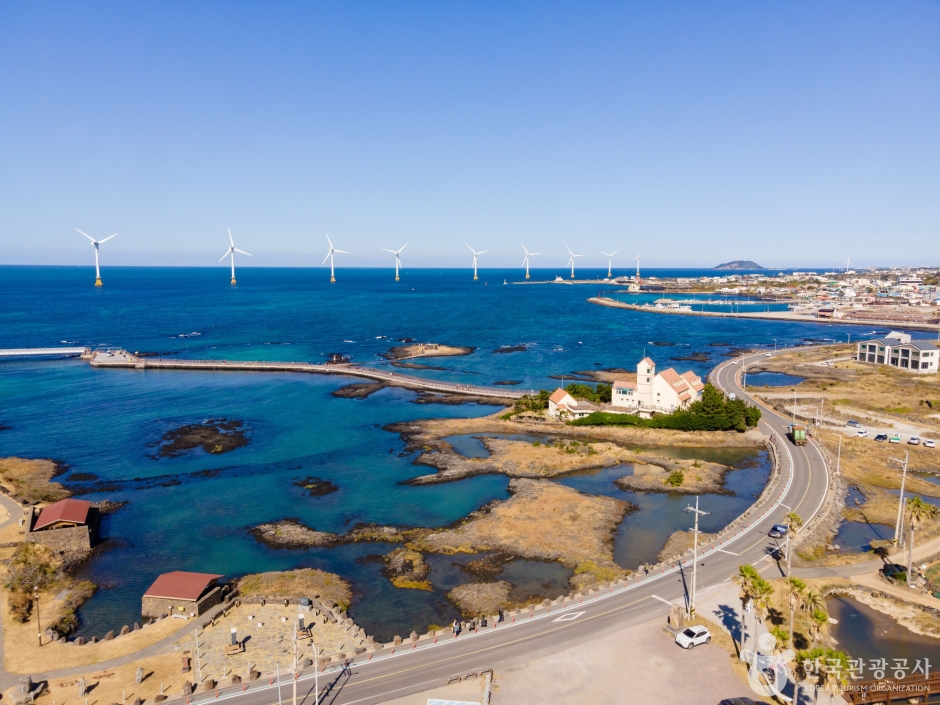 A line of white wind turbines standing in shallow water along the Sinchang coast of Jeju, with a coastal road, black volcanic reefs and clear blue sea