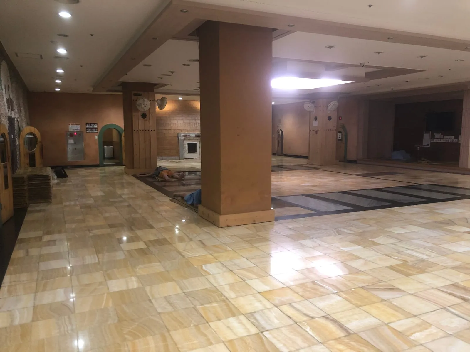 The wide stone-floored common hall of a Korean sauna, with wooden arched doorways to several heated rooms along the walls and a few people asleep on mats on the floor