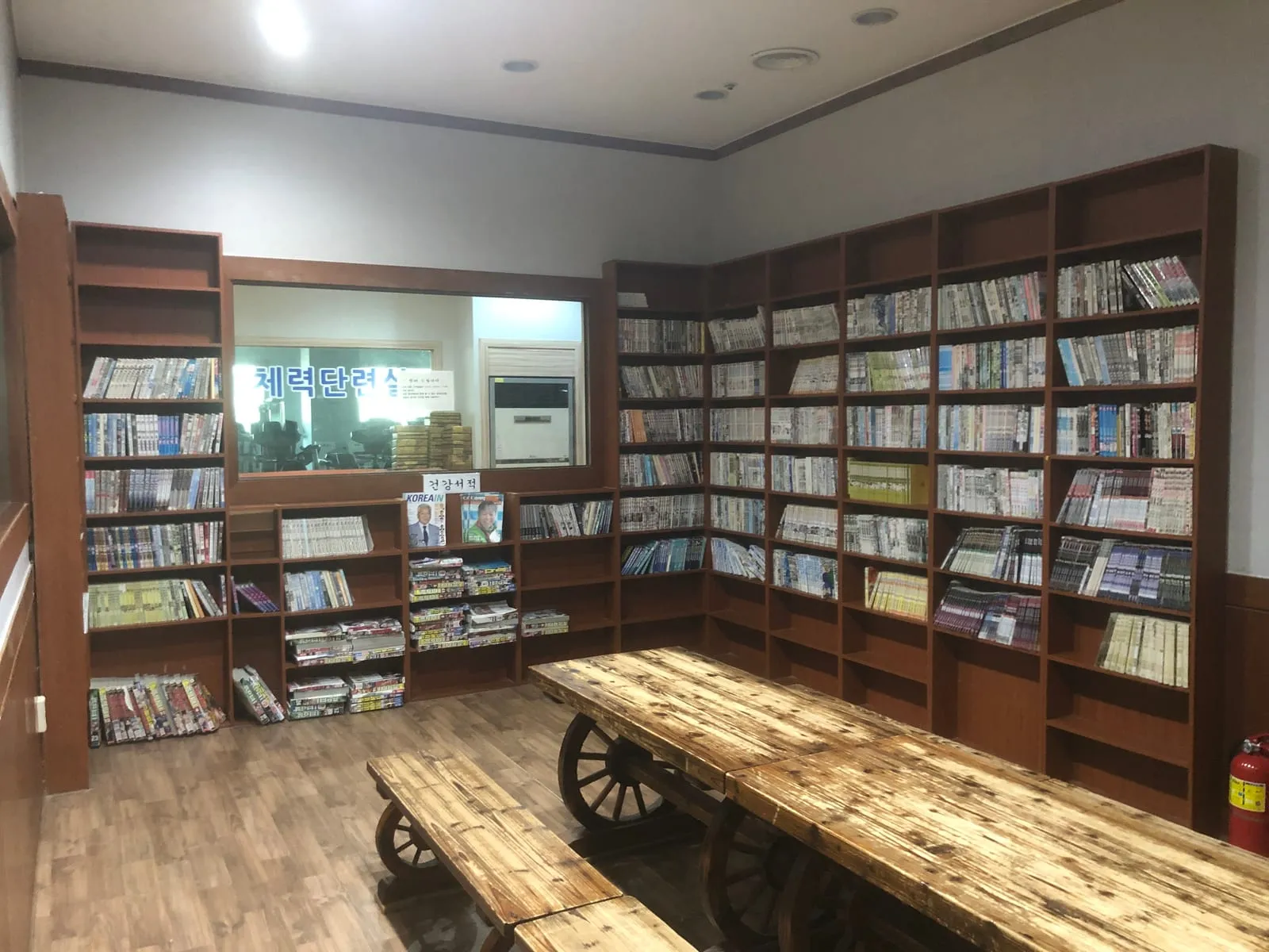A room lined floor to ceiling with shelves of Korean comics, with long wooden benches and a window through to a small gym
