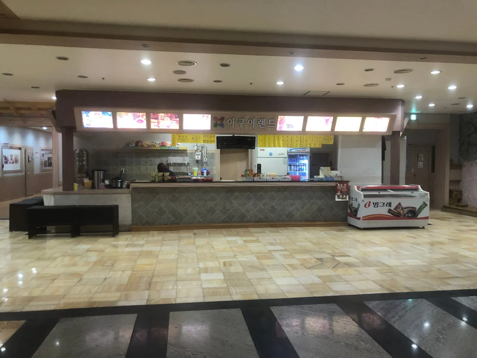 A snack counter inside a jjimjilbang, with trays of eggs and cups of instant noodles on the top, a drinks fridge, an ice-cream freezer and menu boards on the wall