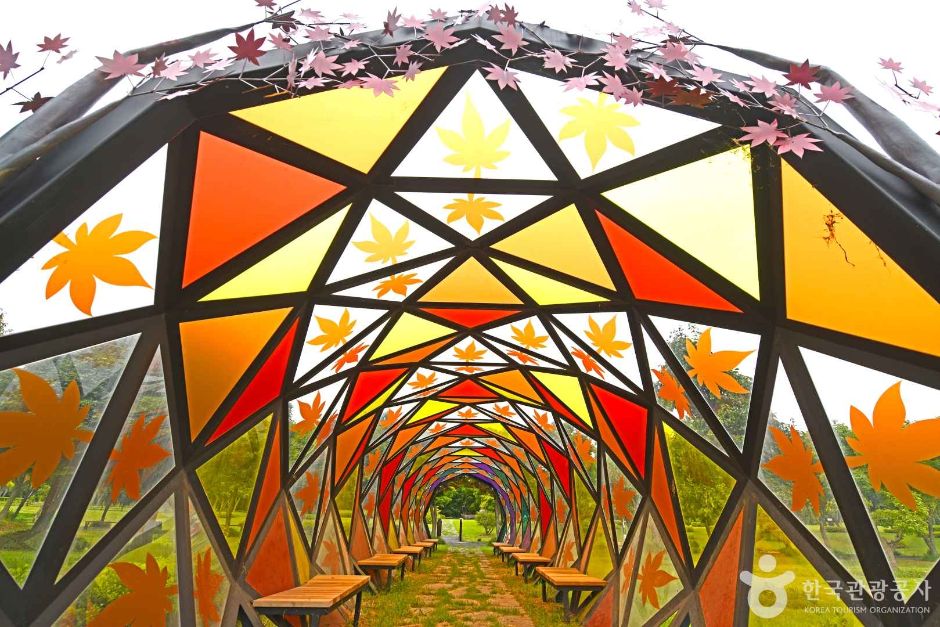 A geodesic dome pavilion glazed with orange and red maple-leaf panels, with a tunnel of maple-leaf glass beyond