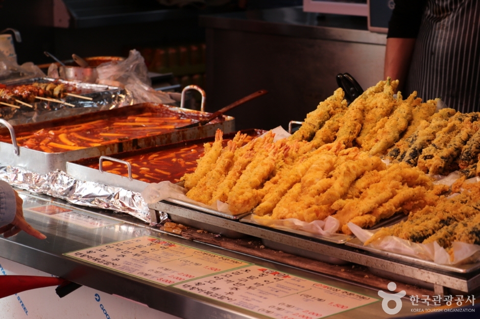Two trays of tteokbokki simmering beside a rack of fried snacks at a Namdaemun Market stall, with a handwritten price list in front