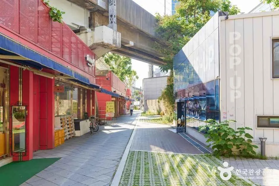 A lane of small shoe shops with red awnings running beneath an elevated concrete viaduct in Seongsu, with large POP UP lettering on the wall opposite
