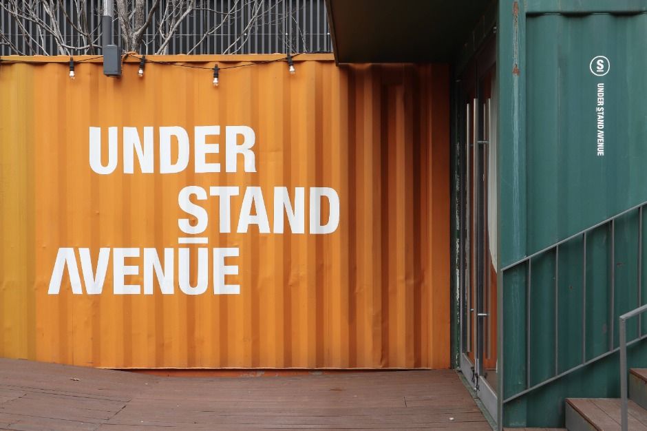 Orange and dark green shipping containers stacked into a building, with UNDER STAND AVENUE painted in white capitals across the orange face