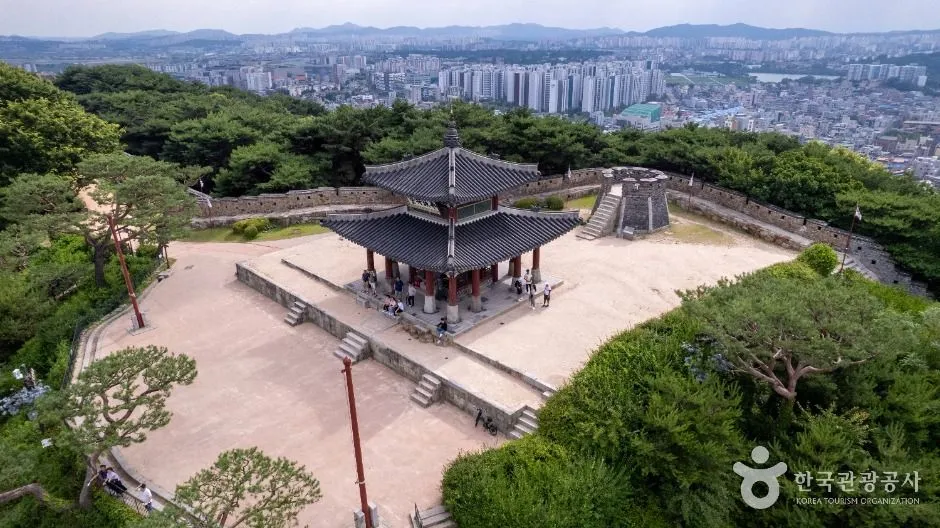 Aerial view of a two-tier pavilion on a broad bare-earth terrace at a hilltop, with the fortress wall running around it and the city spread out below