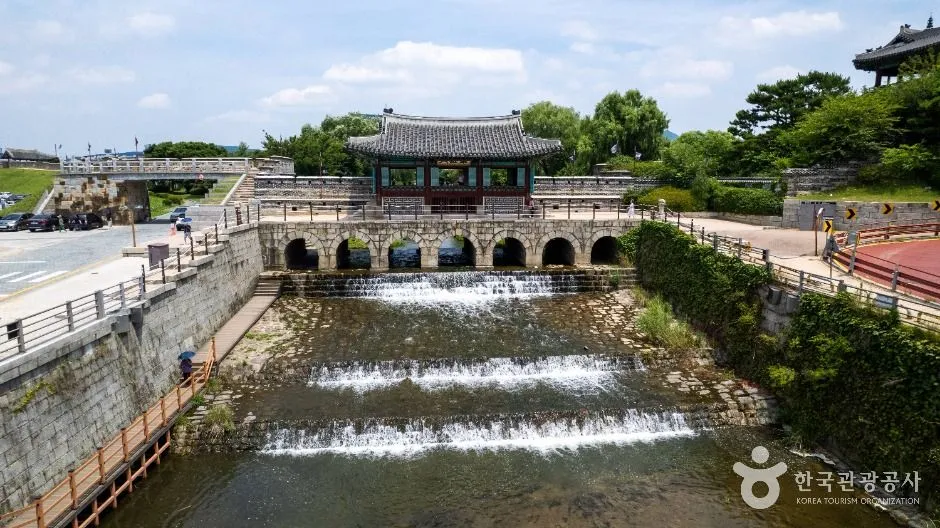 A stone water gate of seven arches spanning a channelled stream, with a tiled wooden pavilion built on top and the water dropping over low weirs below it