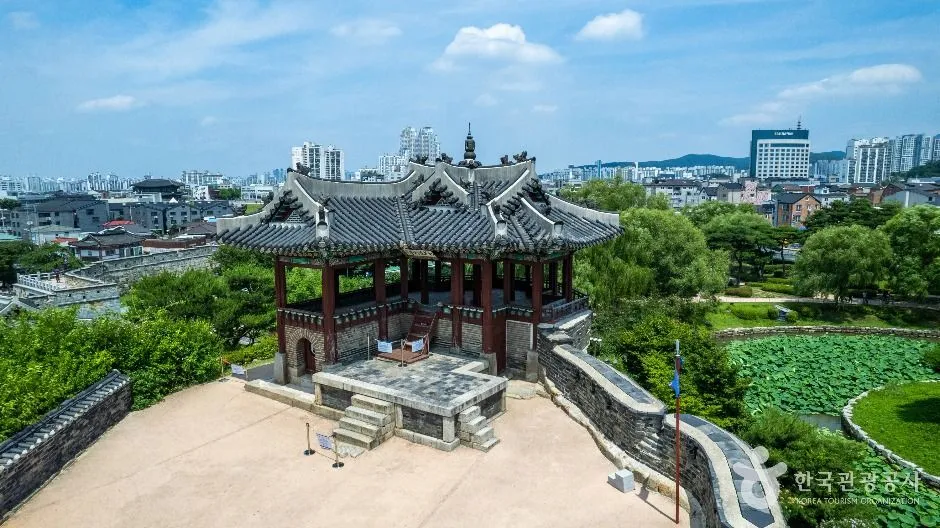 An ornate pavilion with a complex tiled roof standing on a rocky outcrop at a corner of the fortress wall, with the modern city behind